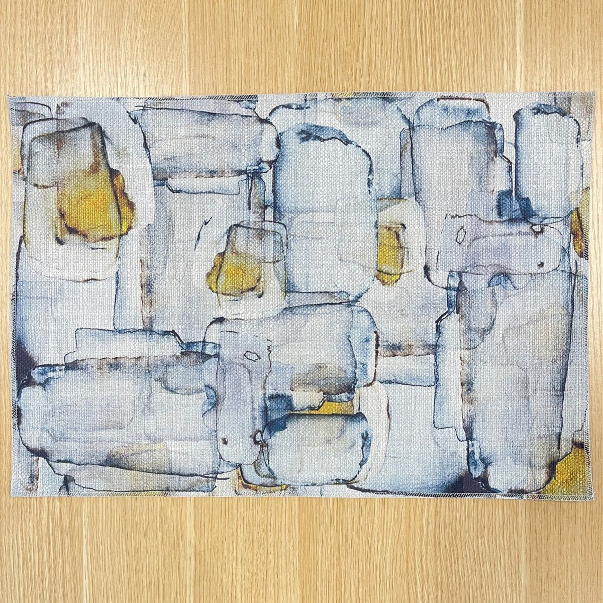Rectangular Mid Century placemat with abstract grey and blue shapes with ochre accents, on a wooden tabletop with gold cutlery.