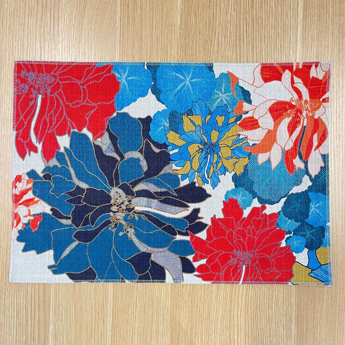Rectangular Sunset placemat with large blue, red and yellow flowers on a pale background, on a wooden tabletop.