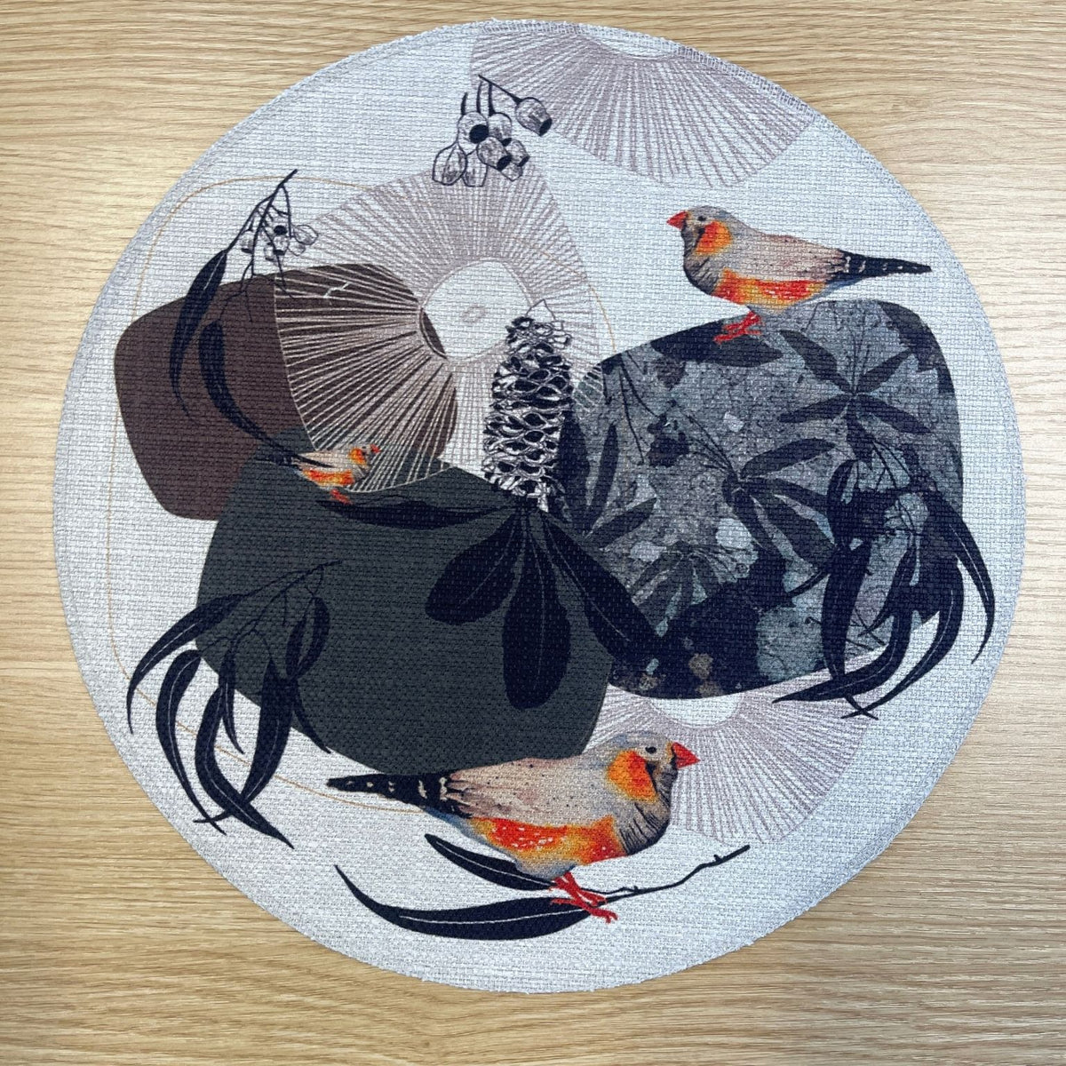 Round Geo Finch placemat with orange-accented finches among black organic shapes and fine geometric lines, on a wooden tabletop.