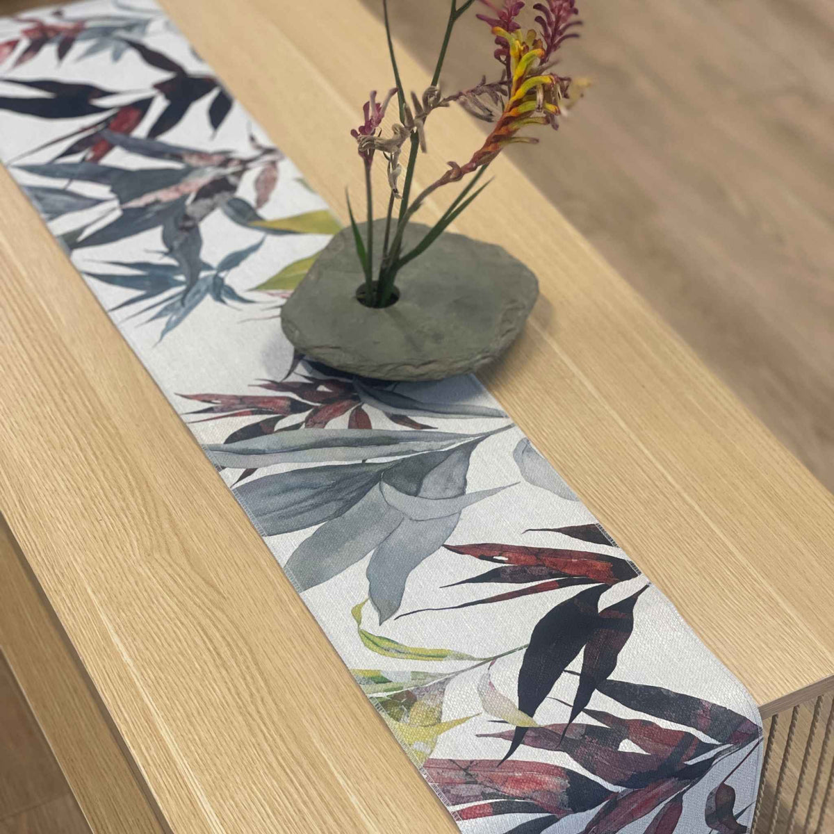 A tropical botanical table runner with a white base and gray, red, and green leaf print, styled on a light wood table with a small stone vase holding dried flowers.