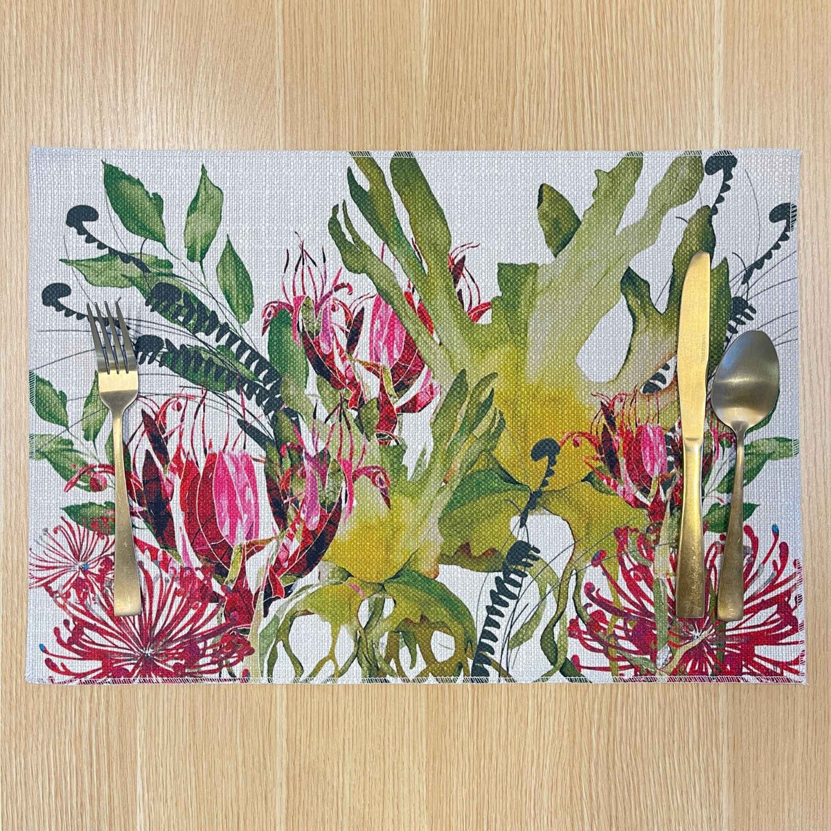 Rectangular Binna Burra placemat with pink-red native flowers among green and yellow foliage, on a wooden tabletop with gold cutlery.