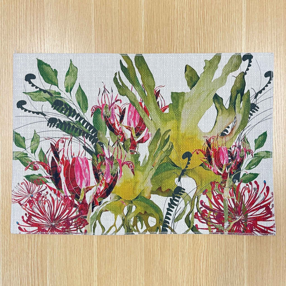 Rectangular Binna Burra placemat with pink-red native flowers among green and yellow foliage, on a wooden tabletop.