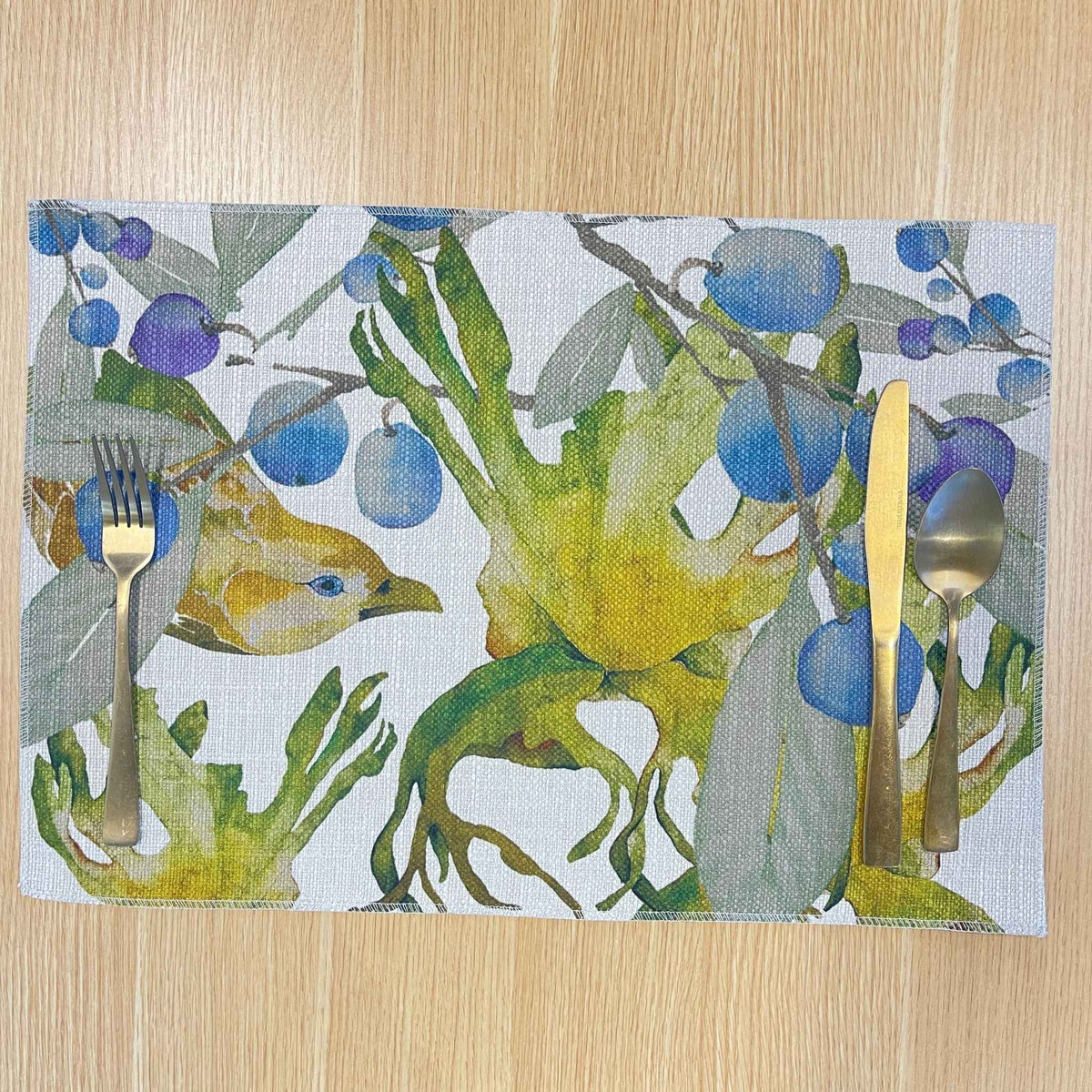 Rectangular Bower placemat with a green bowerbird among blue-purple berries and green foliage, on a wooden tabletop with gold cutlery.