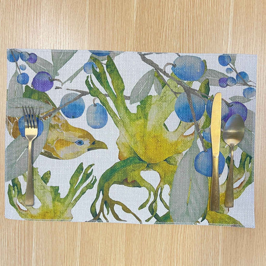 Rectangular Bower placemat with a green bowerbird among blue-purple berries and green foliage, on a wooden tabletop with gold cutlery.