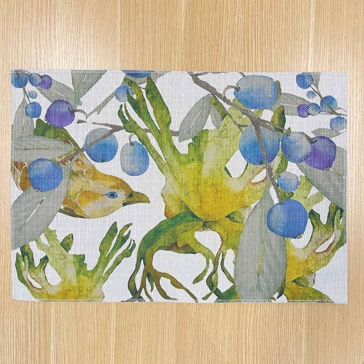 Rectangular Bower placemat with a green bowerbird among blue-purple berries and green foliage, on a wooden tabletop.
