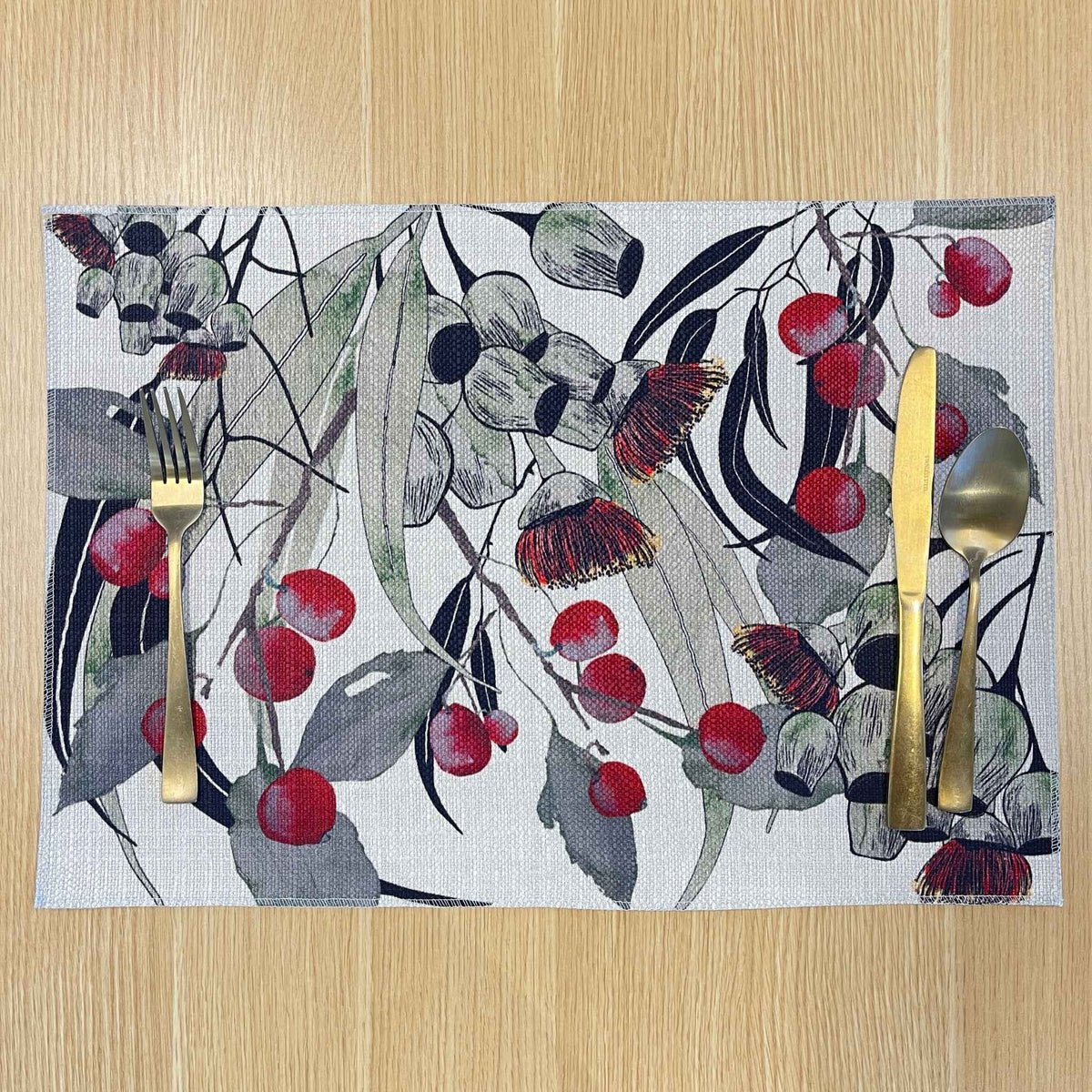 Rectangular Bush Celebration placemat with red gum blossoms and berries among grey-green foliage and dark curved lines, on a wooden tabletop with gold cutlery.