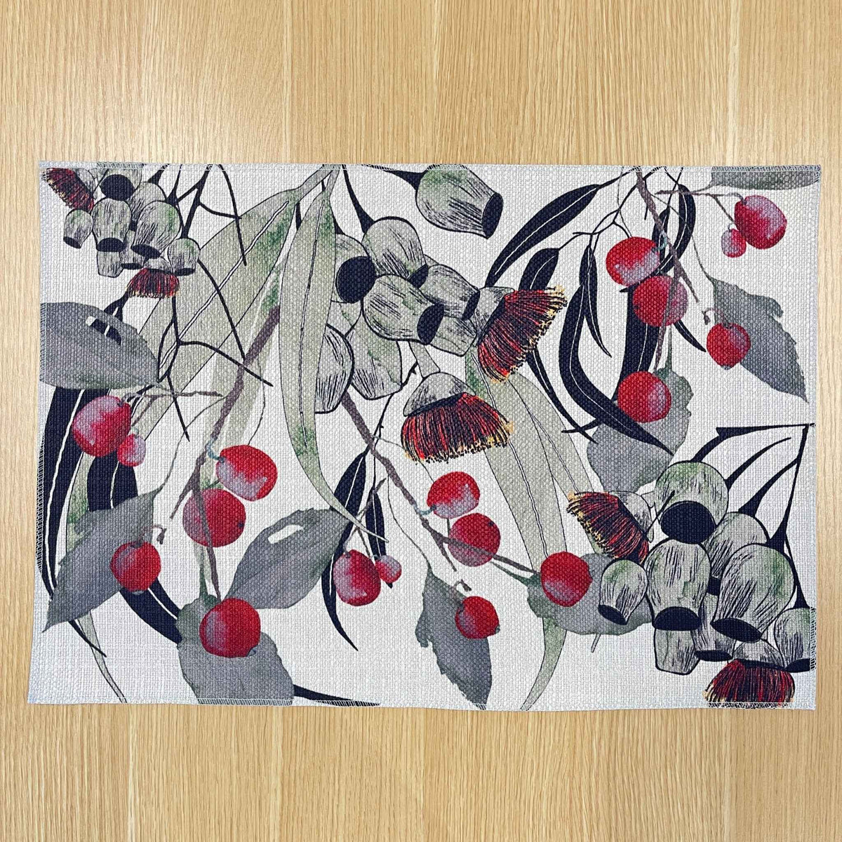 Rectangular Bush Celebration placemat with red gum blossoms and berries among grey-green foliage and dark curved lines, on a wooden tabletop.