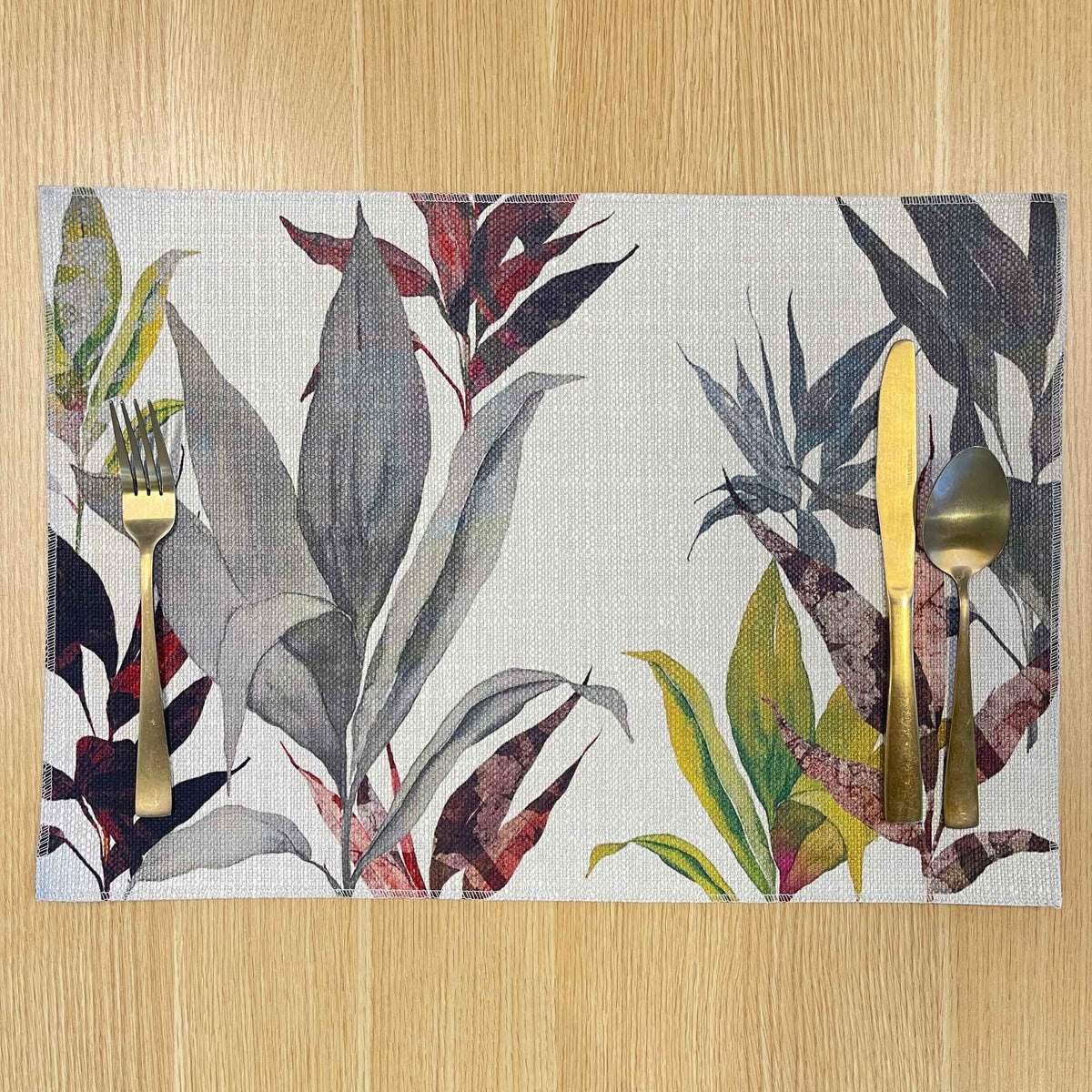 Rectangular Cordyline placemat with layered charcoal, burgundy and olive tropical leaves, on a wooden tabletop with gold cutlery.
