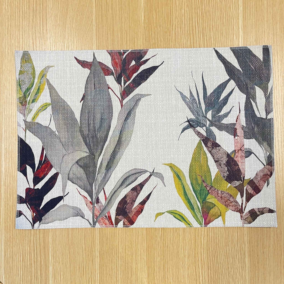 Rectangular Cordyline placemat with layered charcoal, burgundy and olive tropical leaves, on a wooden tabletop.