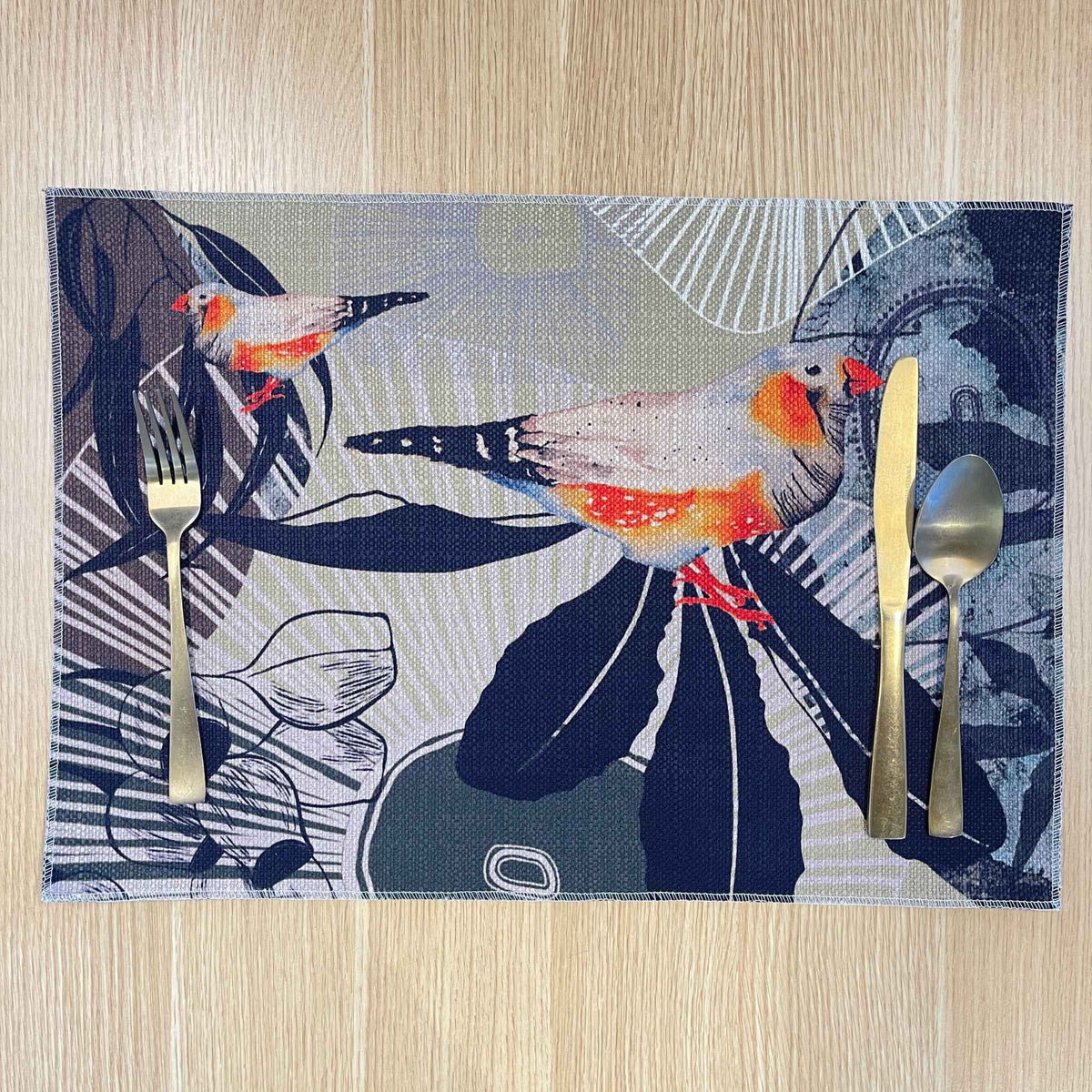 Rectangular Finch Canopy placemat with orange and grey finches among dark leaves and fine botanical lines, on a wooden tabletop with gold cutlery.