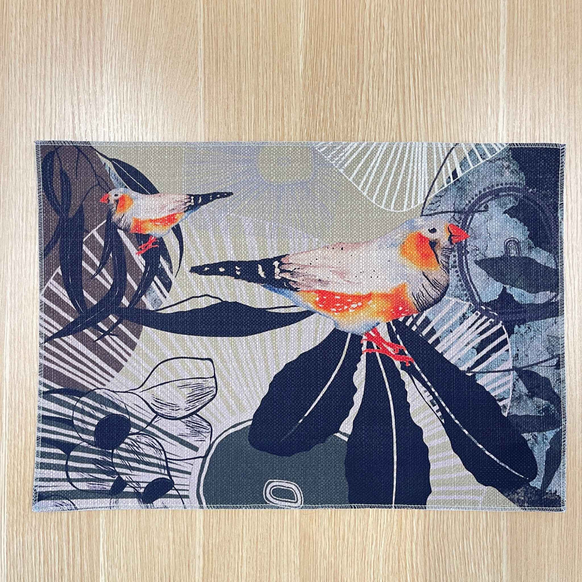 Rectangular Finch Canopy placemat with orange and grey finches among dark leaves and fine botanical lines, on a wooden tabletop.