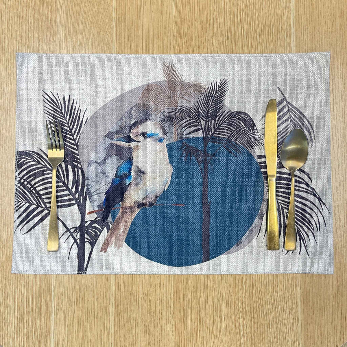 Rectangular Kookaburra placemat with a kookaburra, black palm fronds and a large blue circle, on a wooden tabletop with gold cutlery.