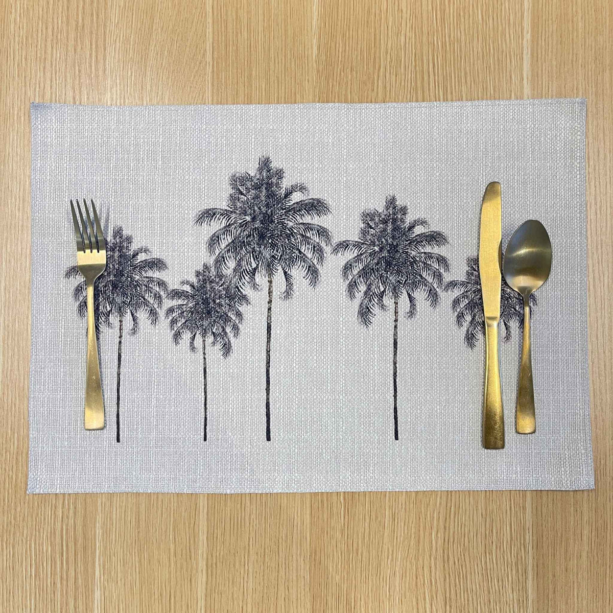 Rectangular Palm Grove placemat with black palm trees on a pale background, on a wooden tabletop with gold cutlery.