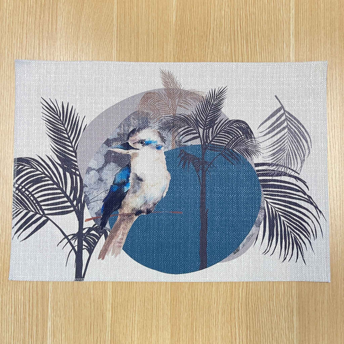 Rectangular Kookaburra placemat with a kookaburra, black palm fronds and a large blue circle, on a wooden tabletop.