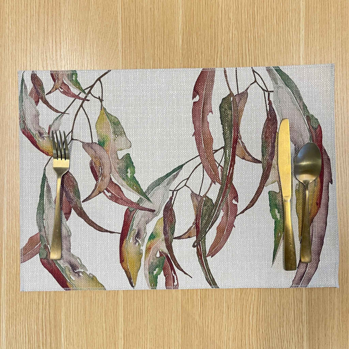 Rectangular Watercolour Gum placemat with watercolour gum leaves in burgundy, green and ochre, on a wooden tabletop with gold cutlery.