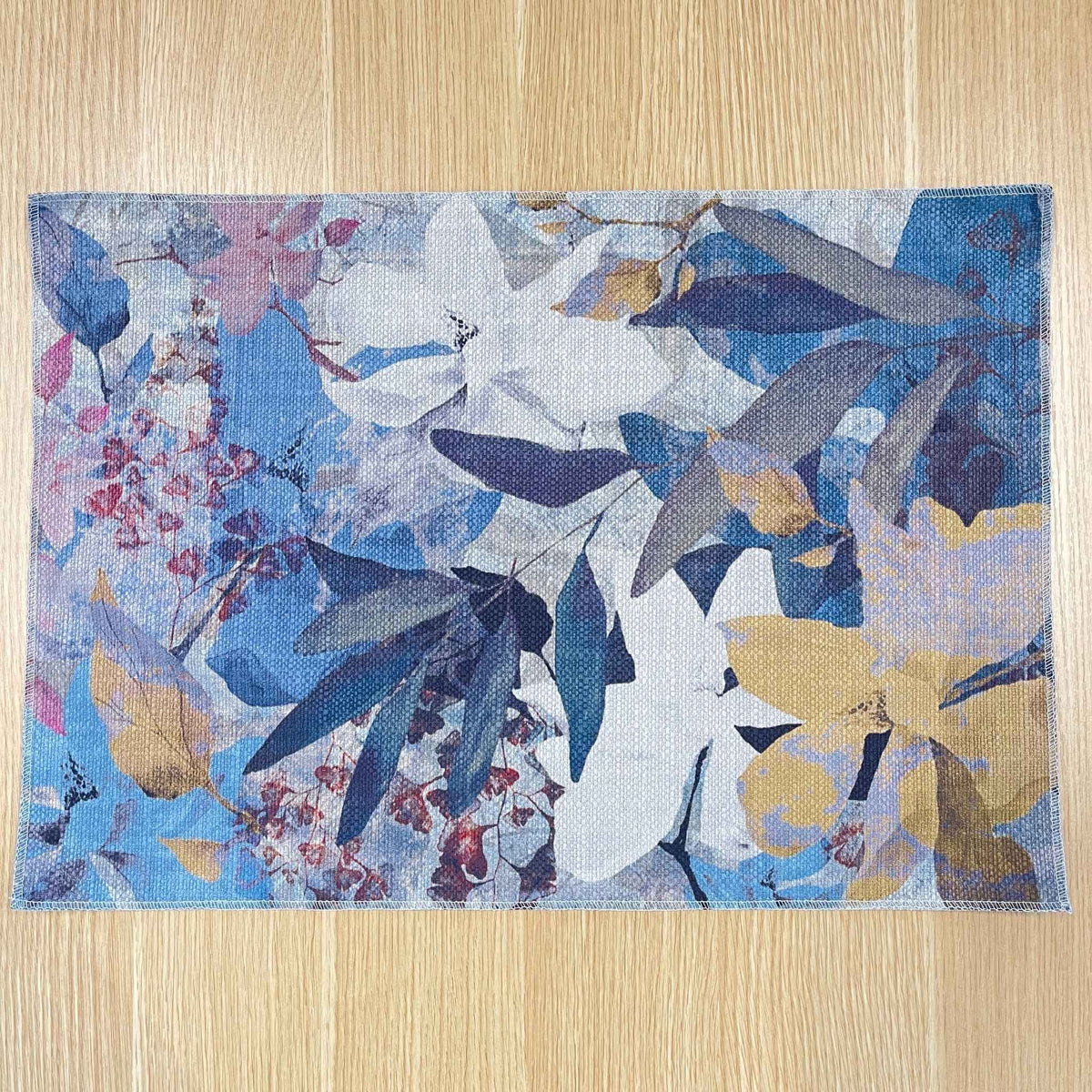 Rectangular Magnolia placemat with layered blue, lavender and white magnolia flowers with ochre accents, on a wooden tabletop.