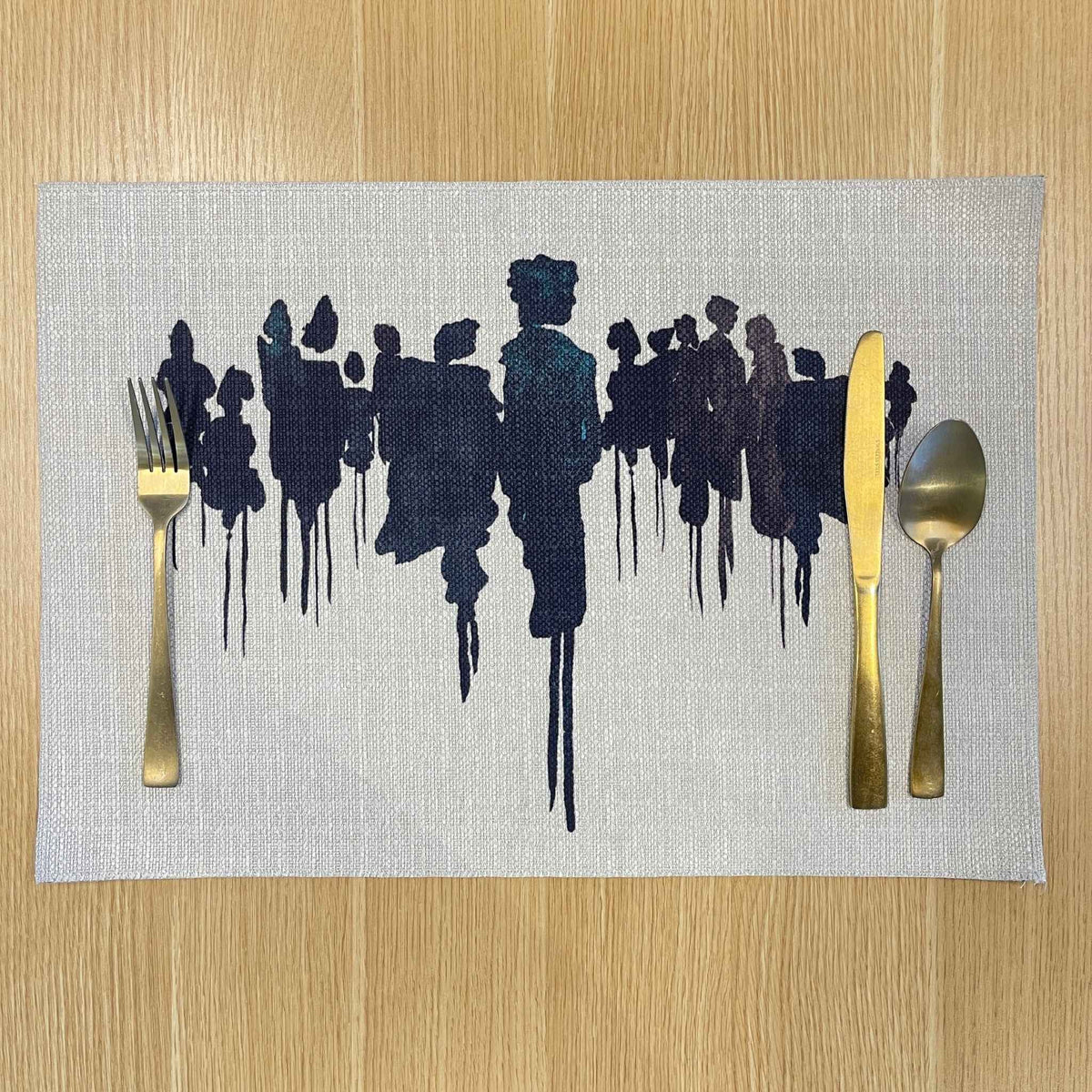 Rectangular Migration placemat with dark abstract silhouettes on a pale background, on a wooden tabletop with gold cutlery.
