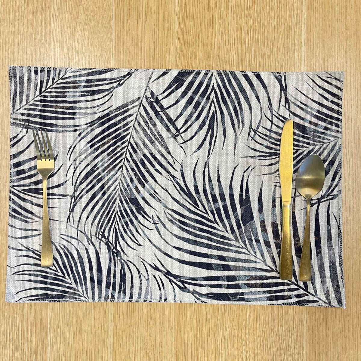 Rectangular Palm Frond placemat with overlapping black and grey palm fronds, on a wooden tabletop with gold cutlery.