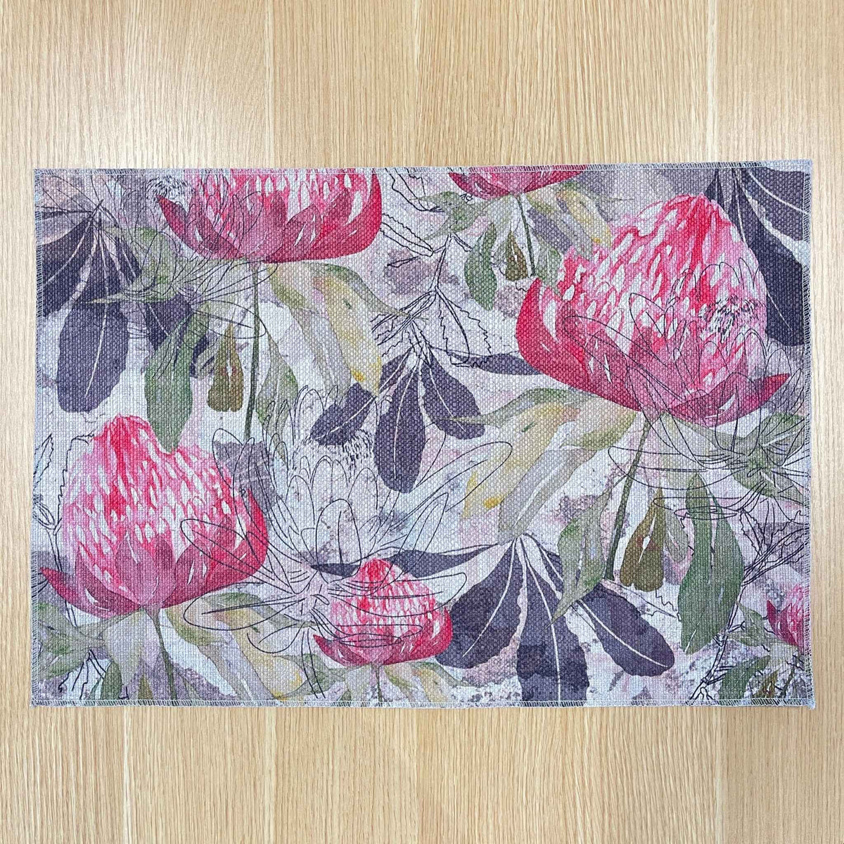 Rectangular Pink Australiana placemat with pink protea flowers among green and grey foliage, on a wooden tabletop with gold cutlery.