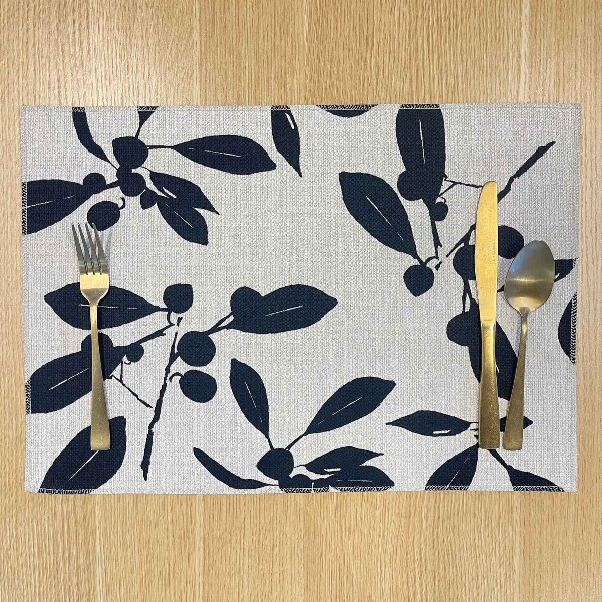 Rectangular Quandong placemat with dark leaf and fruit silhouettes on a pale background, on a wooden tabletop with gold cutlery.