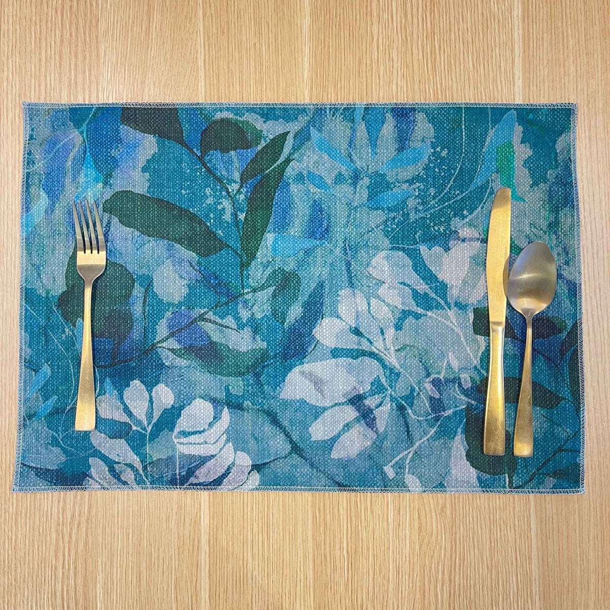 Rectangular Rock Pool placemat with layered turquoise, blue and green foliage, on a wooden tabletop with gold cutlery.