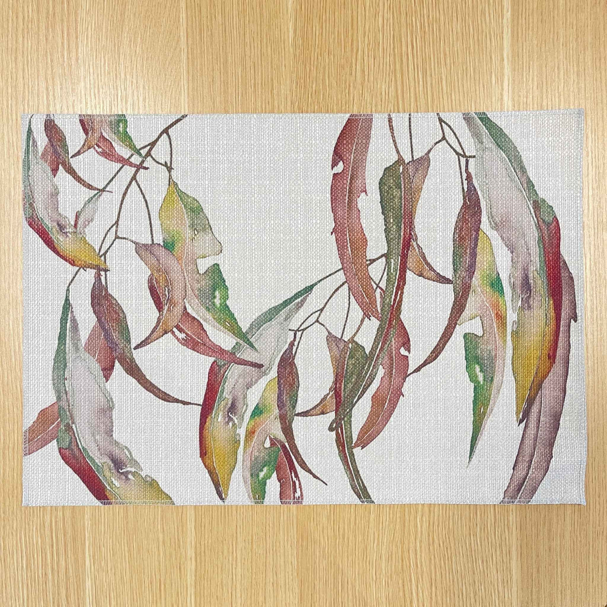 Rectangular Watercolour Gum placemat with watercolour gum leaves in burgundy, green and ochre, on a wooden tabletop.