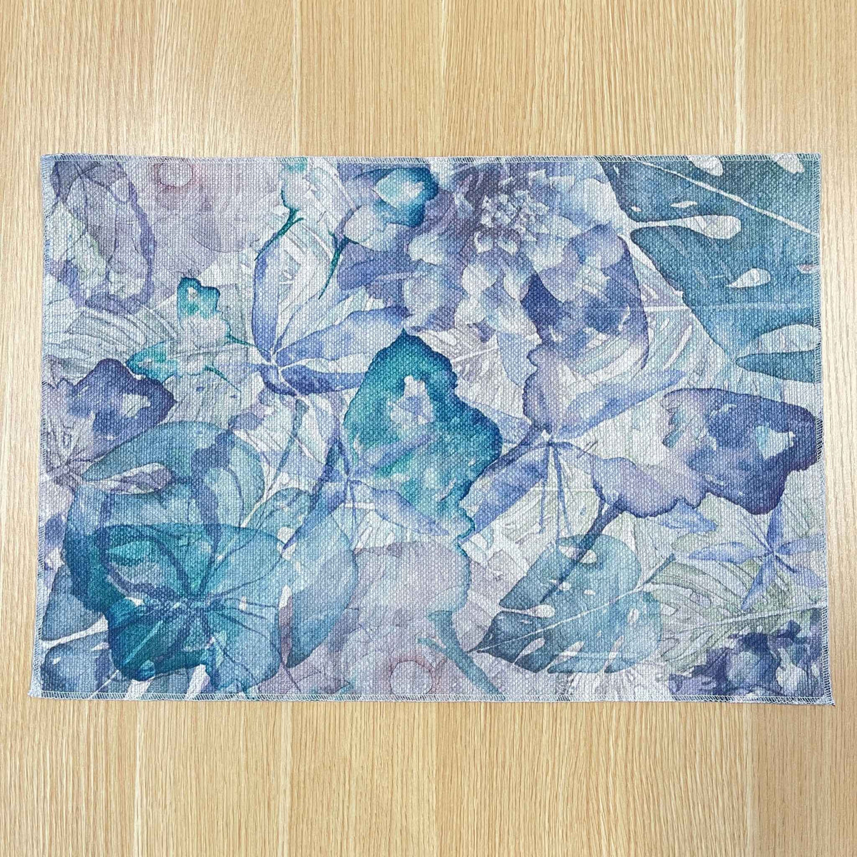 Rectangular Wild Garden placemat with layered blue, lavender and teal botanical shapes, on a wooden tabletop.