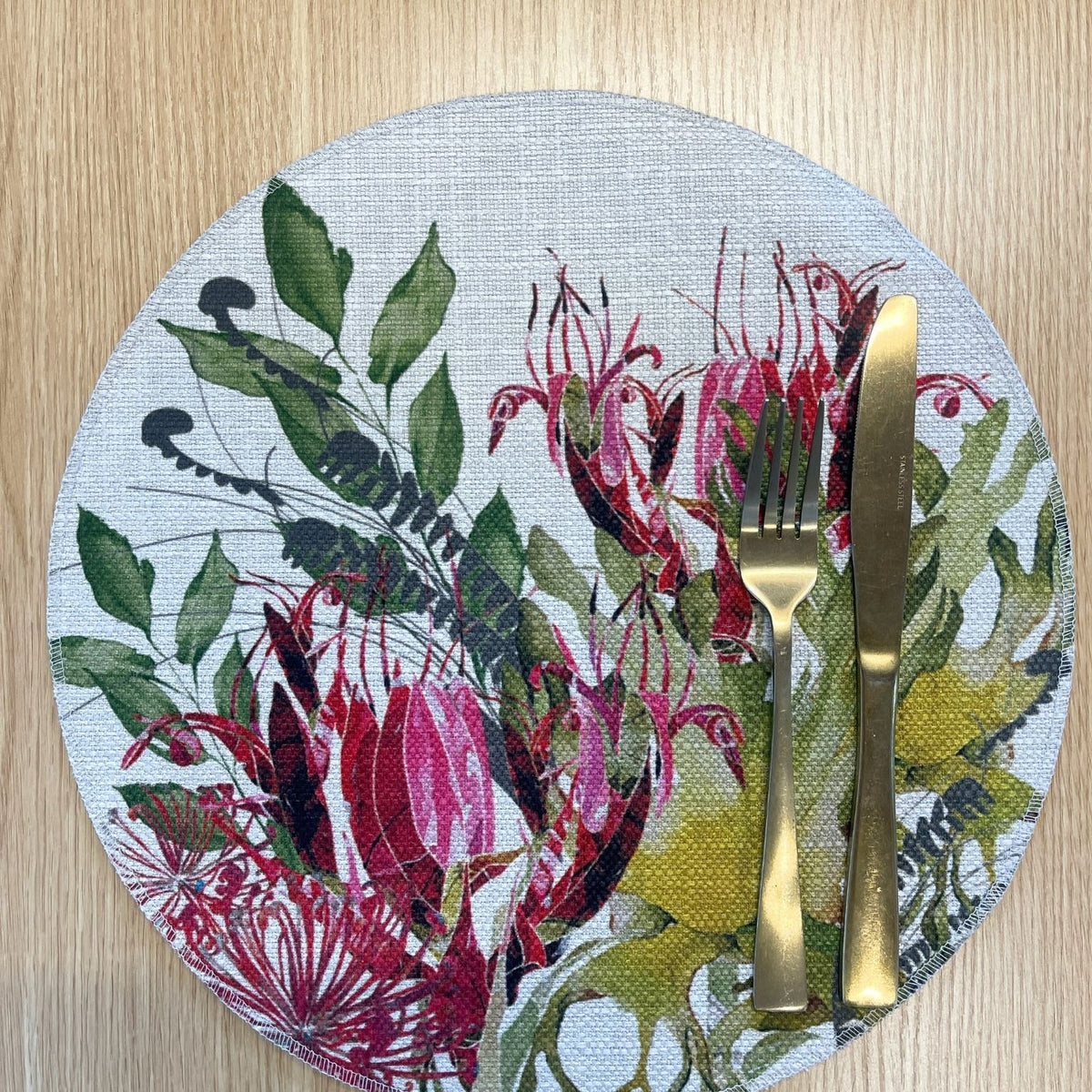 Round Binna Burra placemat with pink-red native flowers among green and yellow foliage, on a wooden tabletop with gold cutlery.