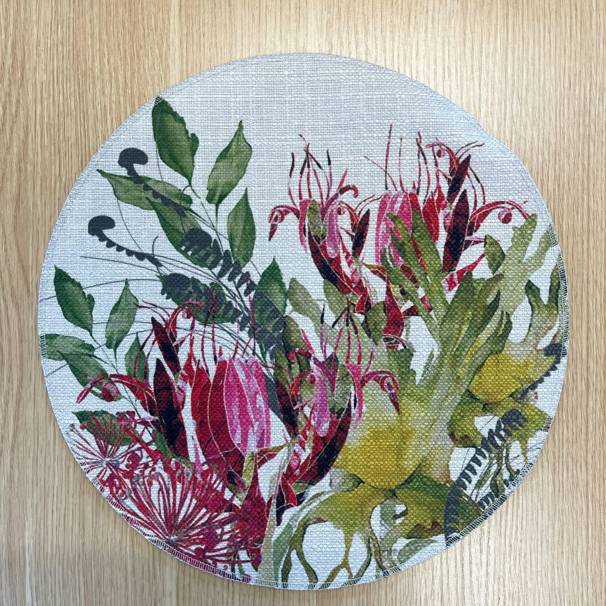Round Binna Burra placemat with pink-red native flowers among green and yellow foliage, on a wooden tabletop.