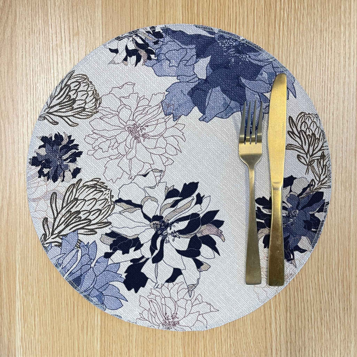 Round Antique Floral Blue placemat with navy and blue flowers with fine floral outlines on a pale background, on a wooden tabletop with gold cutlery.
