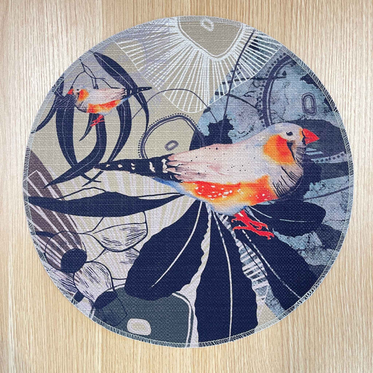 Round Finch Canopy placemat with orange and grey finches among dark leaves and fine botanical lines, on a wooden tabletop.