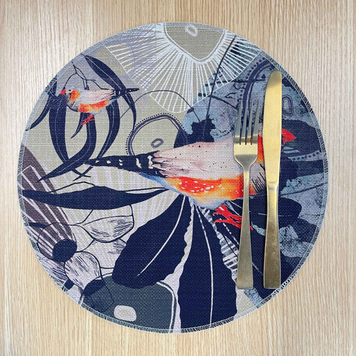Round Finch Canopy placemat with orange and grey finches among dark leaves and fine botanical lines, on a wooden tabletop with gold cutlery.
