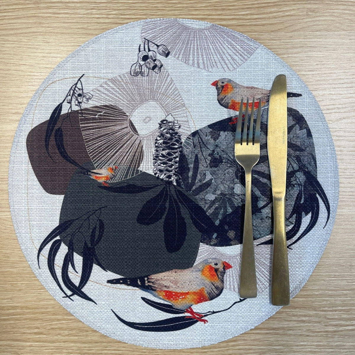 Round Geo Finch placemat with orange-accented finches among black organic shapes and fine geometric lines, on a wooden tabletop with gold cutlery.