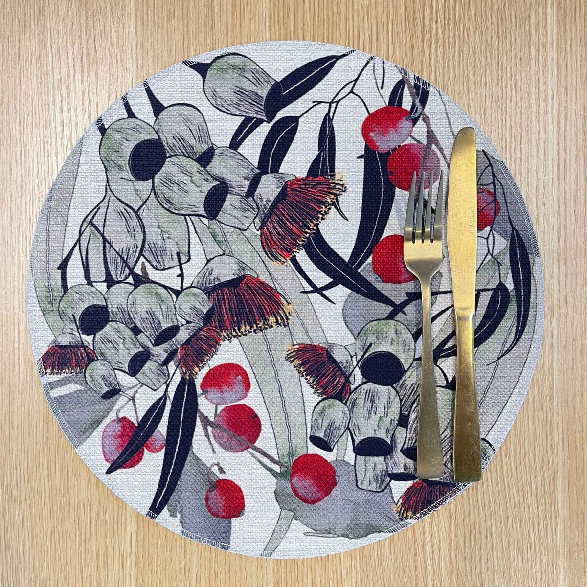 Round Bush Celebration placemat with red gum blossoms and berries among grey-green foliage and dark curved lines, on a wooden tabletop with gold cutlery.