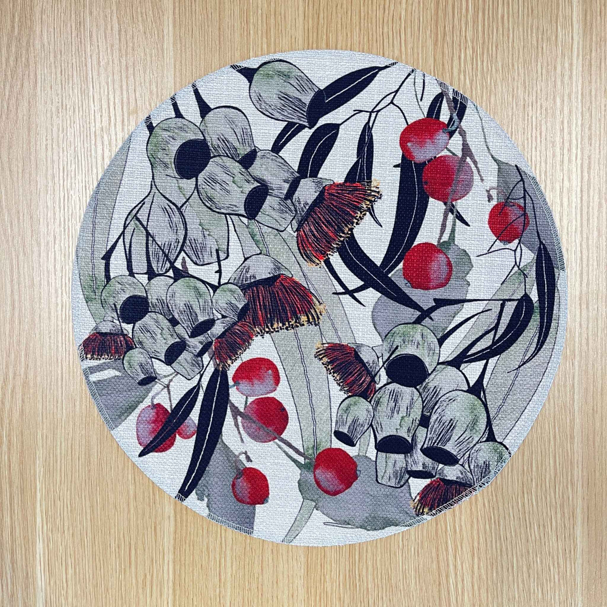 Round Bush Celebration placemat with red gum blossoms and berries among grey-green foliage and dark curved lines, on a wooden tabletop.