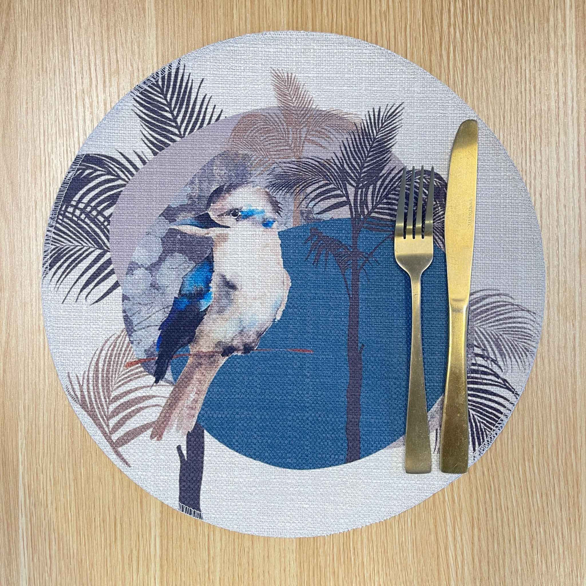 Round Kookaburra placemat with a kookaburra, black palm fronds and a large blue circle, on a wooden tabletop with gold cutlery.