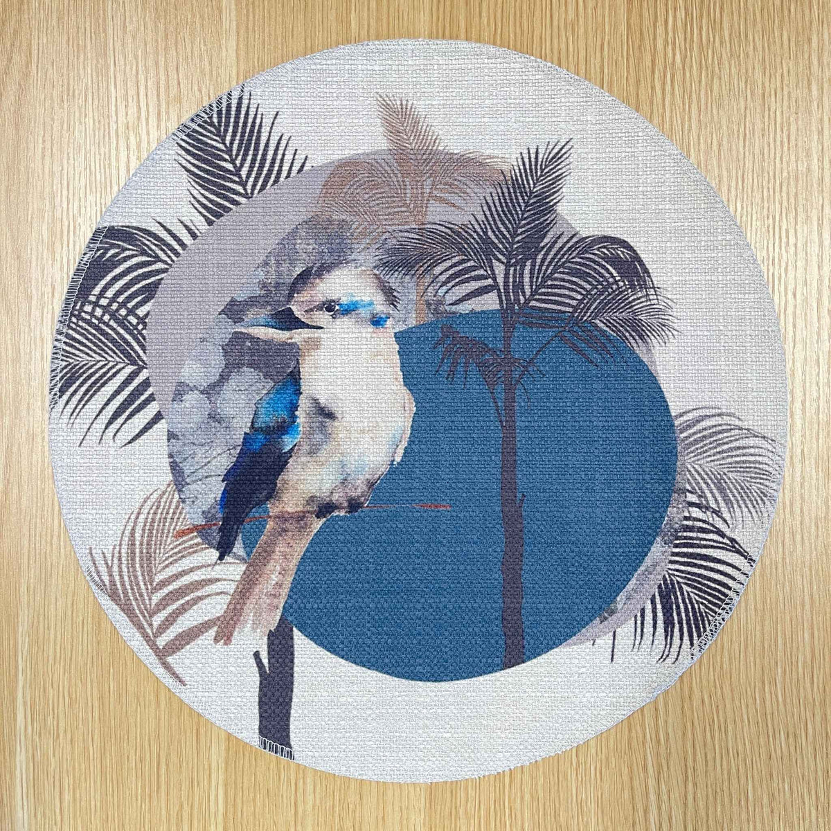 Round Kookaburra placemat with a kookaburra, black palm fronds and a large blue circle, on a wooden tabletop.