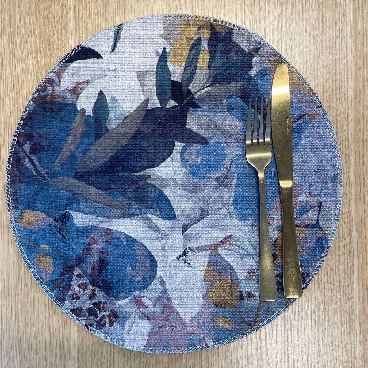 Round Magnolia placemat with layered blue, lavender and white magnolia flowers with ochre accents, on a wooden tabletop with gold cutlery.