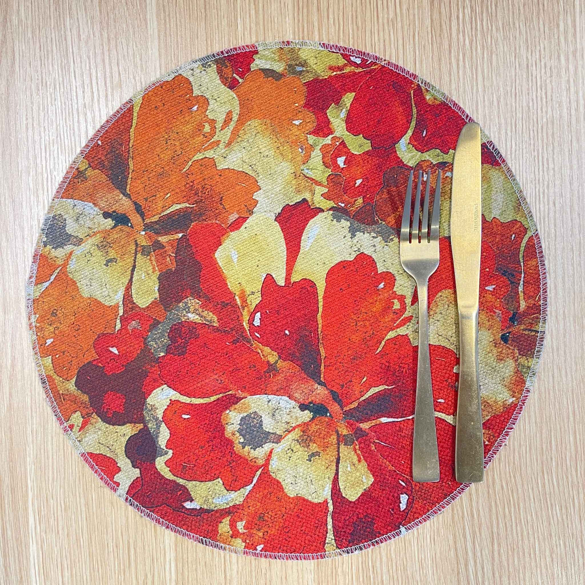 Round Marigold Detail placemat with layered red, orange and gold flowers, on a wooden tabletop with gold cutlery.