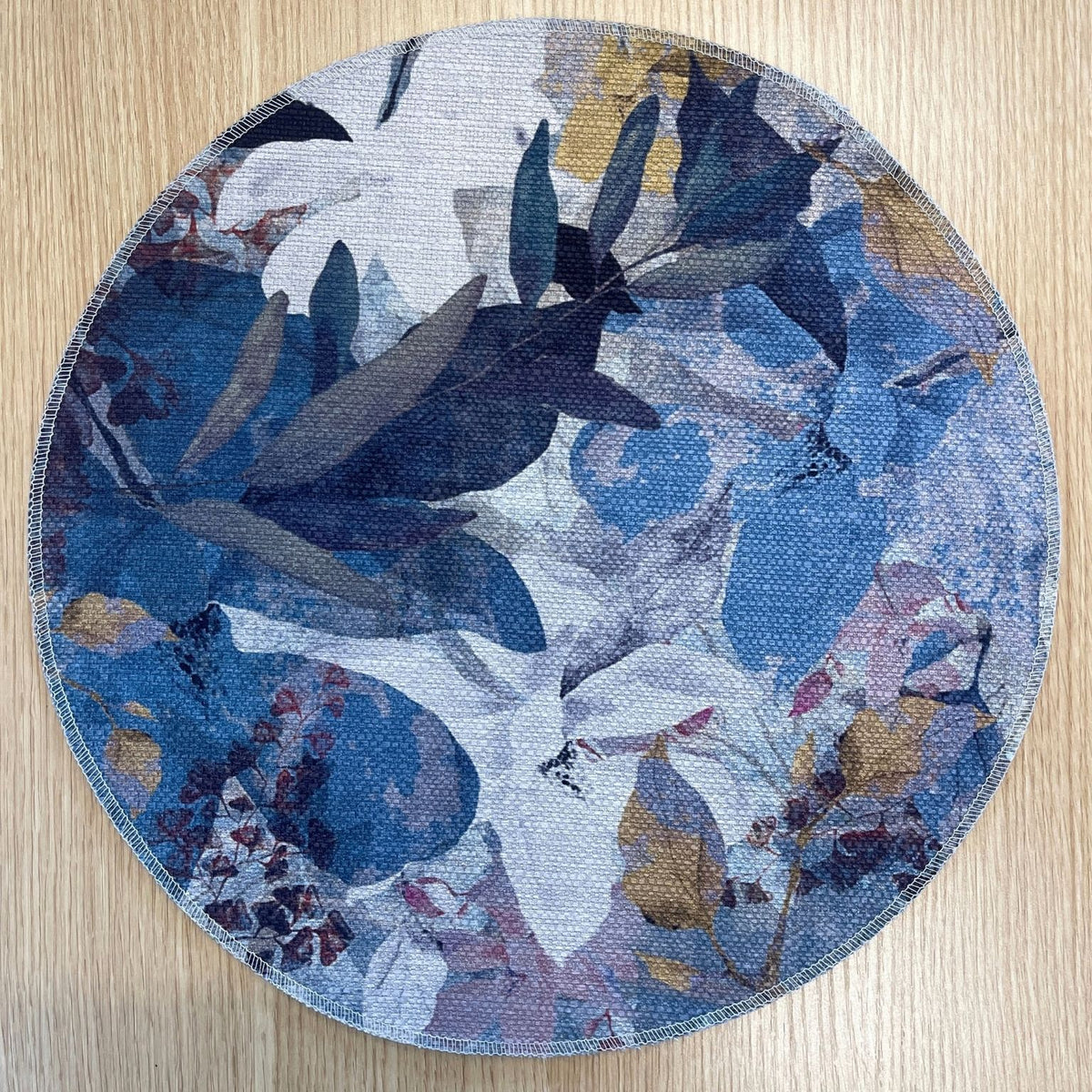 Round Magnolia placemat with layered blue, lavender and white magnolia flowers with ochre accents, on a wooden tabletop.