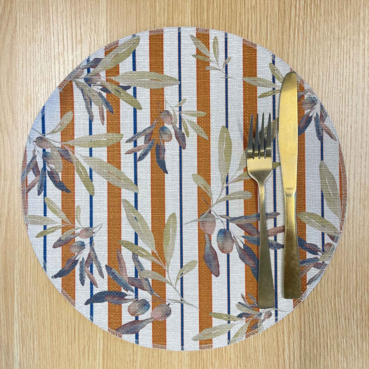 Round Olive and Sol placemat with olive branches over terracotta and navy stripes, on a wooden tabletop with gold cutlery.