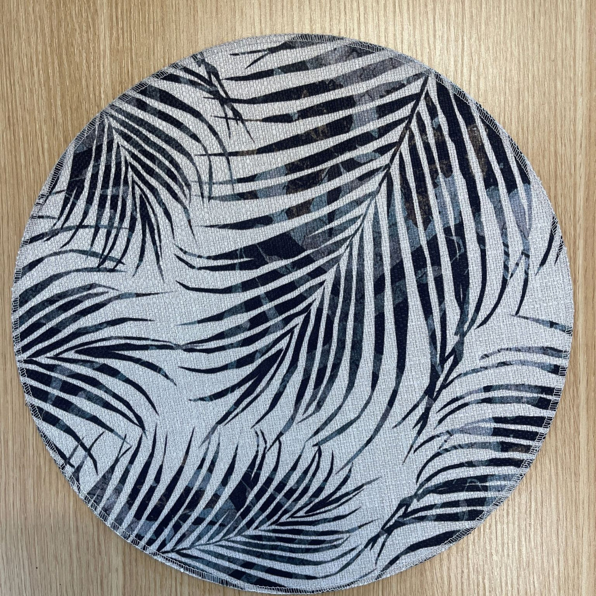 Round Palm Frond placemat with overlapping black and grey palm fronds, on a wooden tabletop.