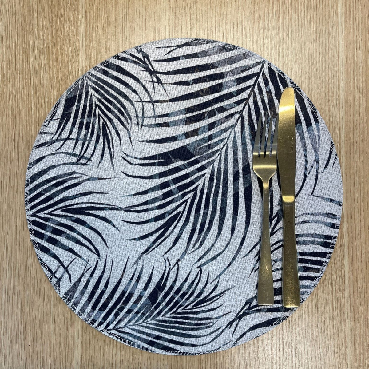 Round Palm Frond placemat with overlapping black and grey palm fronds, on a wooden tabletop with gold cutlery.