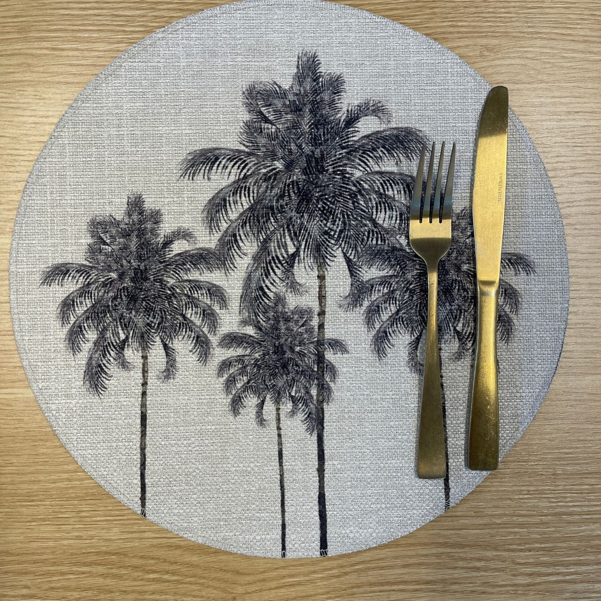 Round Palm Grove placemat with black palm trees on a pale background, on a wooden tabletop with gold cutlery.