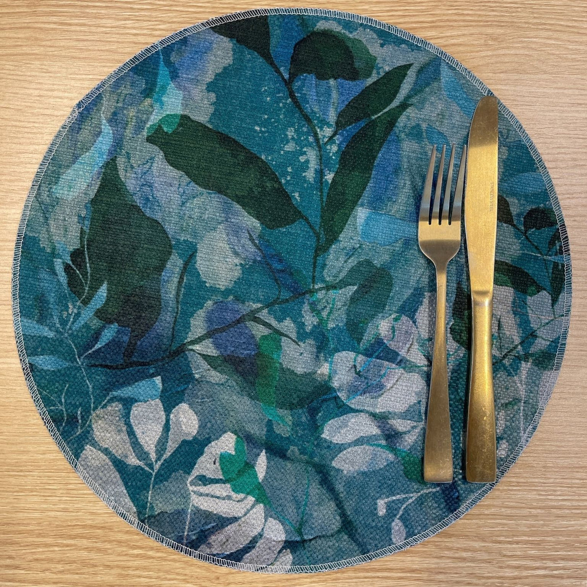 Round Rock Pool placemat with layered turquoise, blue and green foliage, on a wooden tabletop with gold cutlery.