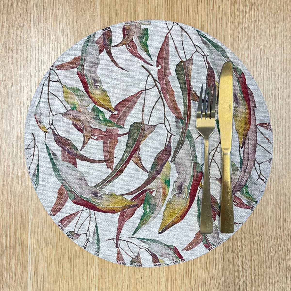 Round Watercolour Gum placemat with watercolour gum leaves in burgundy, green and ochre, on a wooden tabletop with gold cutlery.