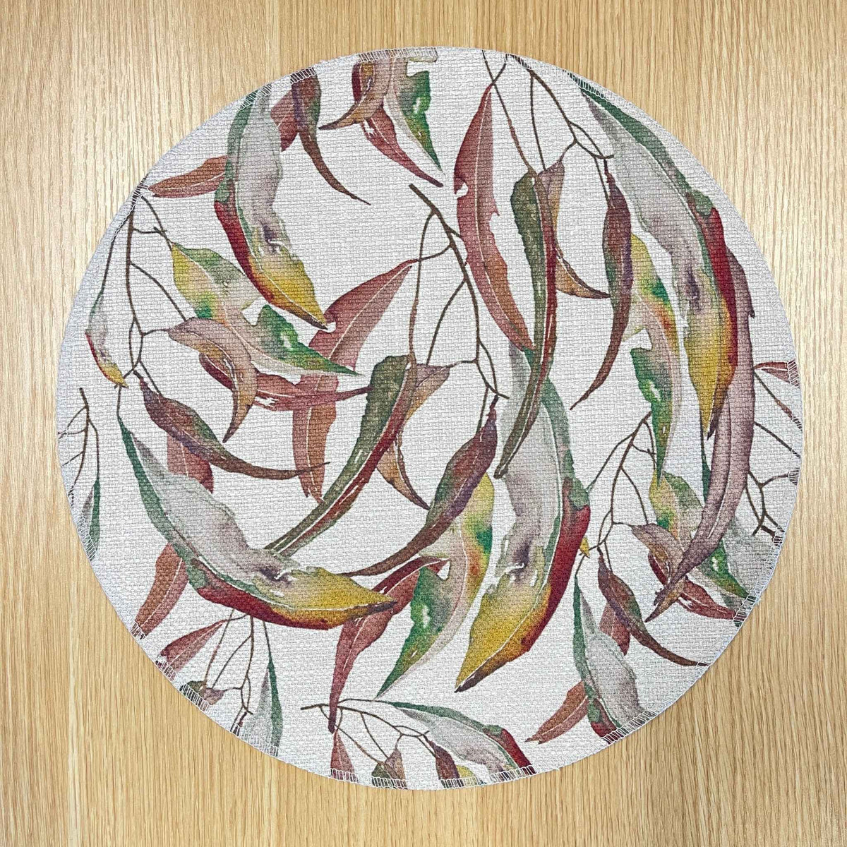 Round Watercolour Gum placemat with watercolour gum leaves in burgundy, green and ochre, on a wooden tabletop.