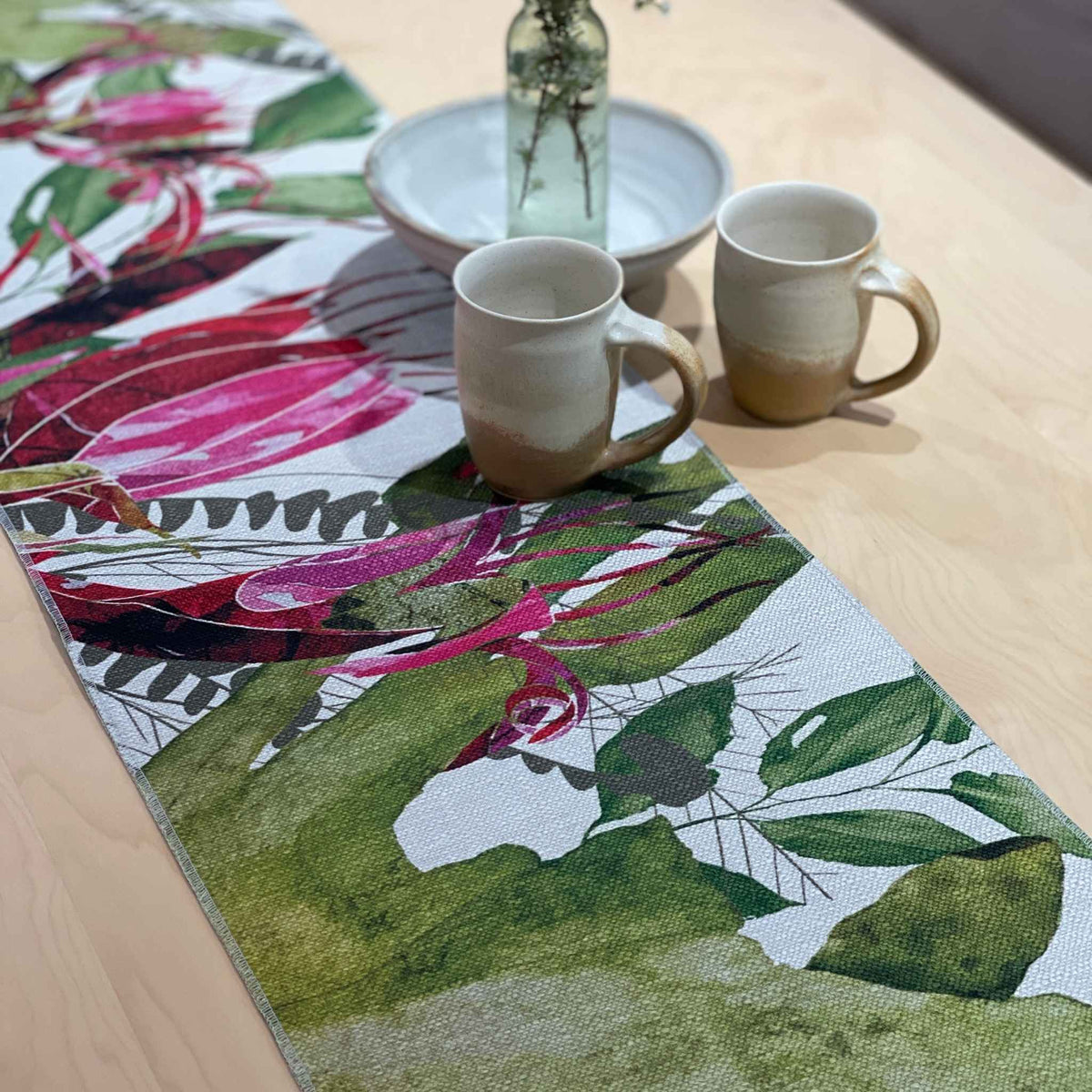 Binna Burra table runner with pink-red native flowers among green and yellow foliage, styled with two cups, a bowl and glass vase