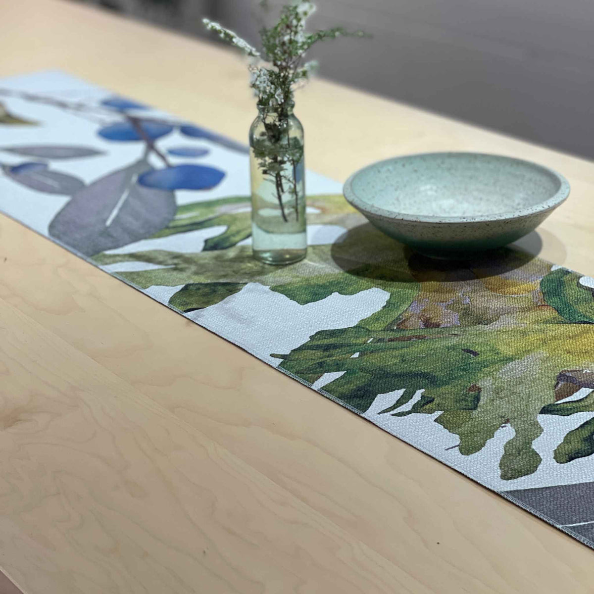 A rectangular woven table runner with a botanical leaf pattern in green, blue, and gray on a white base, styled on a light wood table with a small glass vase and a ceramic bowl.