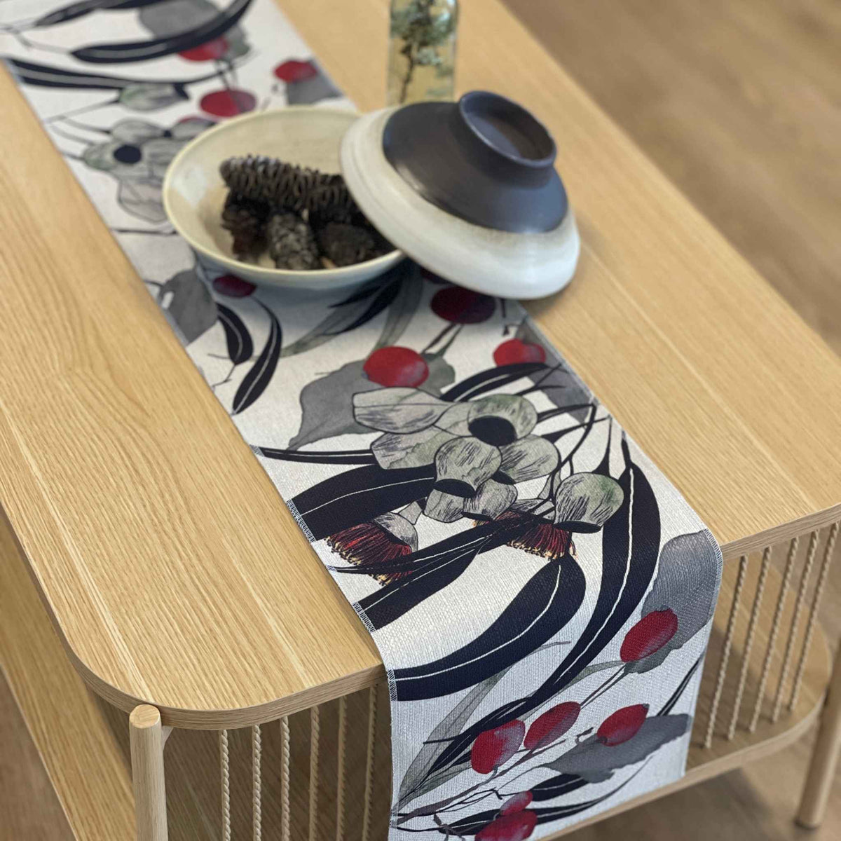 A rectangular table runner with a botanical pattern of gray flowers, dark navy leaves, and red berries, styled on a light wood table with a bowl and a ceramic dish.